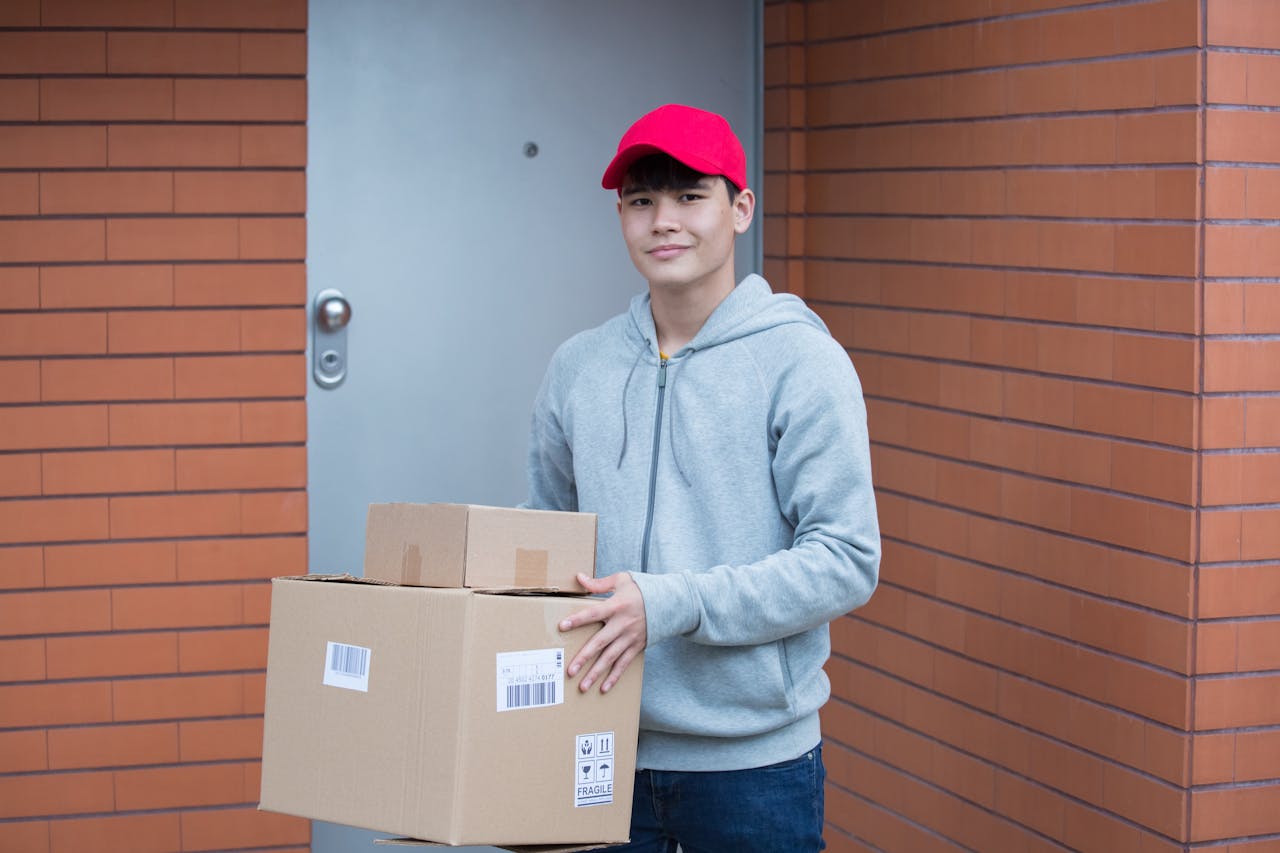 Courier in red cap and gray hoodie delivering packages at a modern brick building.